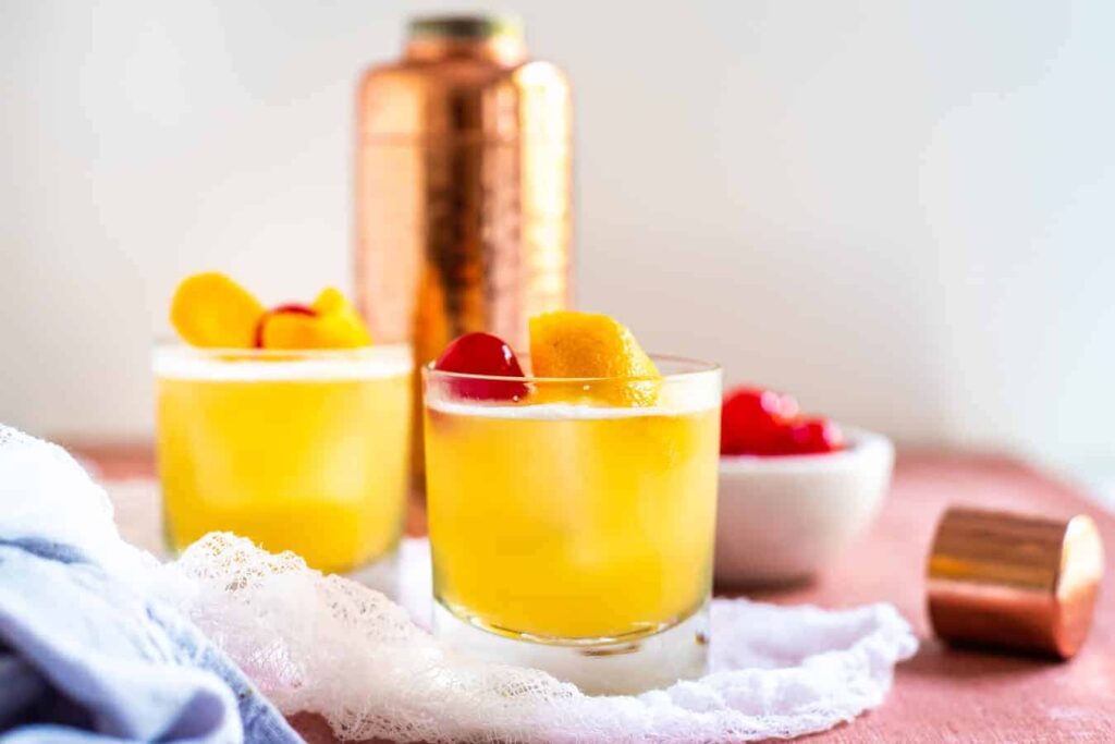 a shaker and two whiskey sour drink on a pink table