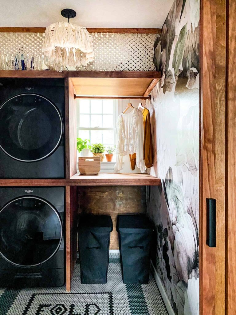 Small Laundry Room makeover. I love the stacked washer and dryer, the dramatic peel and stick wallpaper and penny tiles in this laundry room. The DIY Shelving around washer and dryer is brilliant . And talk about that macrame curtain...! Wow. Such a great laundry room makeover.