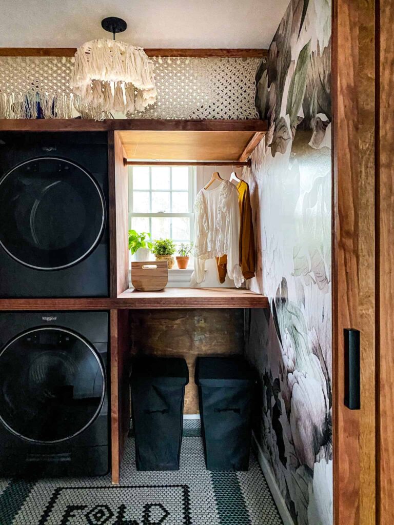 Small Laundry Room makeover. I love the stacked washer and dryer, the dramatic peel and stick wallpaper and penny tiles in this laundry room. The DIY Shelving around washer and dryer is brilliant . And talk about that macrame curtain...! Wow. Such a great laundry room makeover.